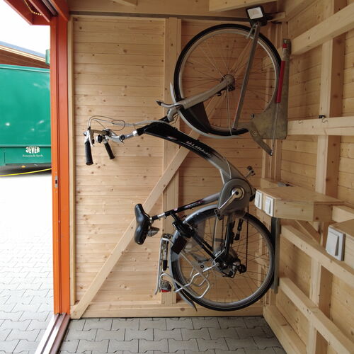 Close-up interior view of a modern timber storage building featuring wall-mounted bike racks that securely hold two bicycles in a vertical position, maximizing storage space. The natural wood interior walls and shelving offer a durable and organized environment for various items. The bright orange frame and sliding gray doors provide a stylish and functional exterior contrast. Designed and installed by Forever Timber and supplied by Bertsch HolzBau, this storage solution is ideal for efficiently storing bicycles and equipment in a compact and visually appealing space.