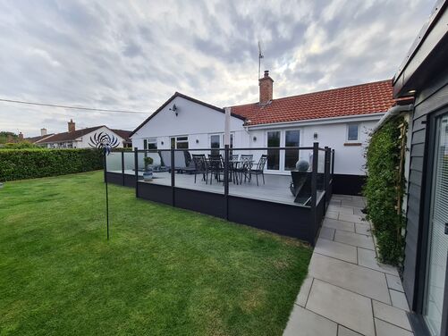 This image showcases a beautifully landscaped backyard with a raised deck that serves as a focal point. The deck, framed with sleek black glass balustrades, adds a contemporary touch, contrasting elegantly with the white facade of the house and the vibrant green lawn. The outdoor dining setup on the deck makes it a perfect space for entertaining or enjoying meals outdoors.

The clean, linear paving stones lead to the side of the deck, seamlessly integrating functionality and style. The neat lawn is well-maintained, and the garden ornament, possibly a kinetic wind spinner, adds an artistic and dynamic feature to the space.

Overall, this backyard is a perfect blend of modern design and natural beauty, creating a welcoming and aesthetically pleasing outdoor area.
