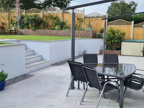 Stylish outdoor patio with large modern grey tiles, complemented by sleek black garden furniture. The multi-tiered design features white retaining walls and neatly landscaped greenery, creating a functional yet elegant space for relaxation and entertaining