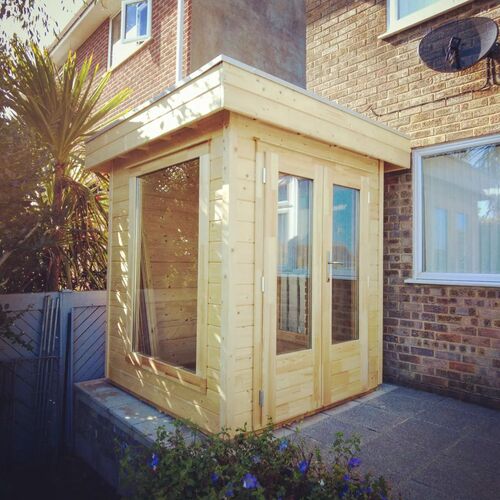 Compact timber garden building with a flat roof, designed to fit snugly against the exterior wall of a brick home. Featuring large glass panels and double doors, the structure offers plenty of natural light and a cozy yet functional interior. The natural wood finish complements the surrounding garden space, while the raised platform with paved flooring adds a neat and practical touch. Ideal as a small home office, reading nook, or relaxation space, this building, designed and installed by Forever Timber, showcases efficient use of space and high-quality craftsmanship
