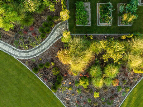 Stunning aerial view of a landscaped garden showcasing a harmonious blend of lush greenery, ornamental plants, and structured vegetable beds. Curved stone pathways wind through the vibrant space, creating a seamless connection between manicured lawns and colorful garden beds, highlighting an elegant and well-planned design.