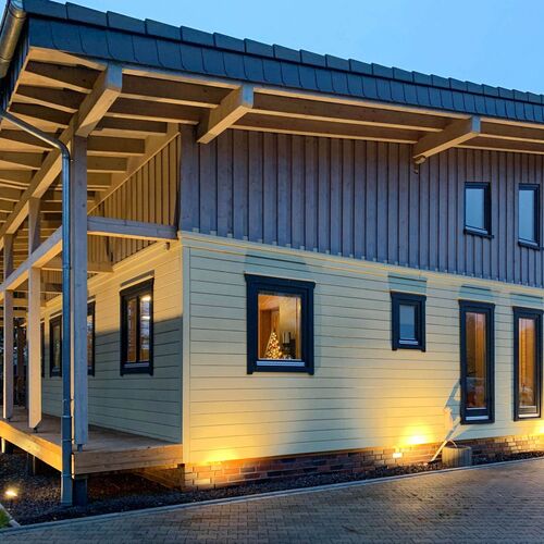 Elegant two-story timber house with a pitched roof, illuminated beautifully at night to showcase its architectural details. The exterior features a combination of vertical and horizontal wooden cladding in natural tones, with dark-framed windows adding a contemporary contrast. Warm interior lighting glows through the large windows, creating a cozy and inviting ambiance. The house includes a covered porch area and is set on a paved driveway, enhancing both functionality and aesthetics. Designed and installed by Forever Timber, this home combines modern design with exceptional craftsmanship, offering both style and comfort.