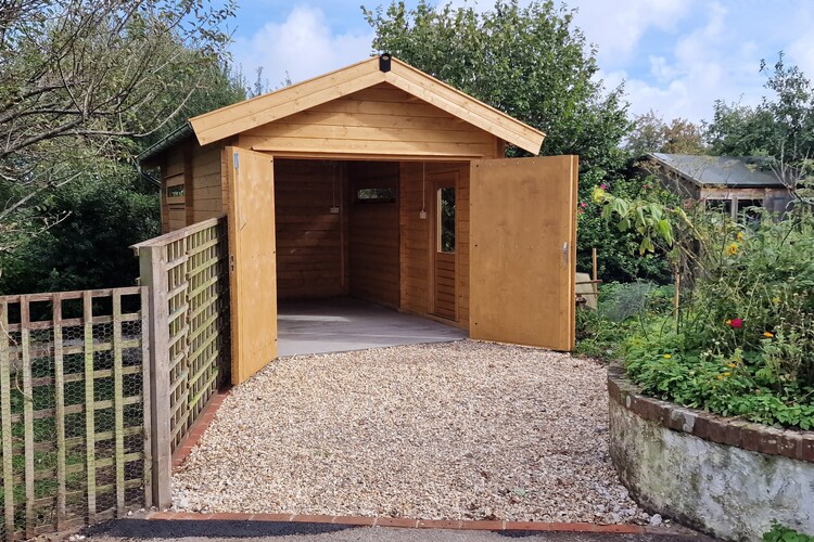 A bespoke wooden garden shed with open double doors is surrounded by greenery. The Lugarde garage sits on a gravel path with a wooden gate to the left and a flower bed with blooming plants to the right, all under a cloudy sky.
