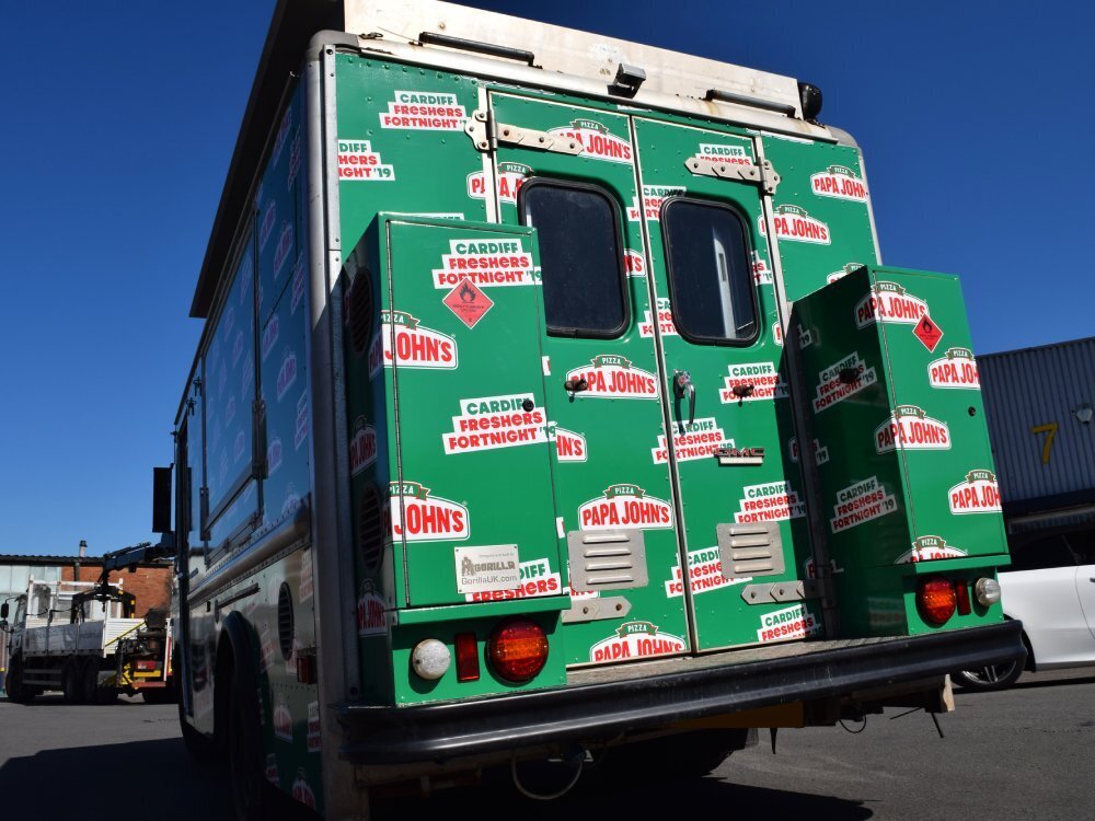 Rear of a GMC truck covered in a green Papa Johns pizza vehicle livery and graphics.