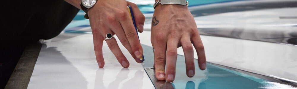 Motorsport vehicle wrap technician cutting high-performance cast vinyl film to size before installation onto race car.