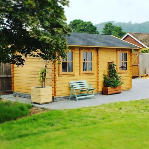 "Charming timber garden room with a pitched roof, featuring large paneled windows and decorative trim that allow natural light to flood the interior. Constructed from premium wood with a natural finish, the structure sits on a solid brick base and is surrounded by a beautifully landscaped area. A cozy outdoor setup includes a pastel green bench and planter boxes, adding a touch of relaxation and style. Supplied by Lugarde and expertly designed and installed by Forever Timber, this garden room serves as a versatile, elegant addition to any outdoor space."