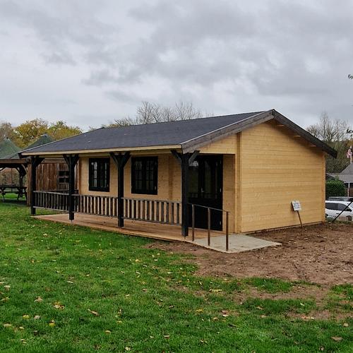 Charming single-story timber cabin with a pitched roof covered in durable shingles. The exterior showcases a natural wood finish, complemented by black-framed windows and doors for a striking contrast. A spacious covered porch runs along the front of the cabin, supported by decorative black timber posts and railings, offering a cozy outdoor seating area. Surrounded by a green lawn, this cabin is ideal for residential or recreational use. Designed and installed by Forever Timber, it blends rustic charm with modern craftsmanship to create a functional and visually appealing structure.