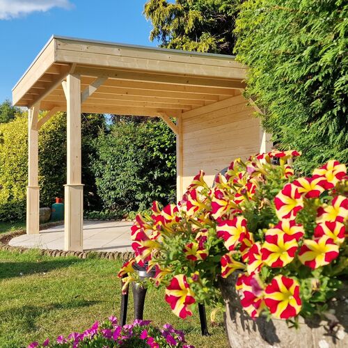 A bespoke garden gazebo supplied by Lugarde and designed and installed by Forevertimber. This elegant structure features a natural timber finish with a flat roof, supported by sturdy wooden posts with decorative bracing. The partially enclosed design includes a solid back wall for privacy and protection from the elements. The gazebo is set on a circular paved base, surrounded by lush greenery, with vibrant red and yellow flowers in the foreground adding a touch of color. This versatile outdoor structure is ideal for relaxation, dining, or entertaining, seamlessly blending with its natural garden surroundings.