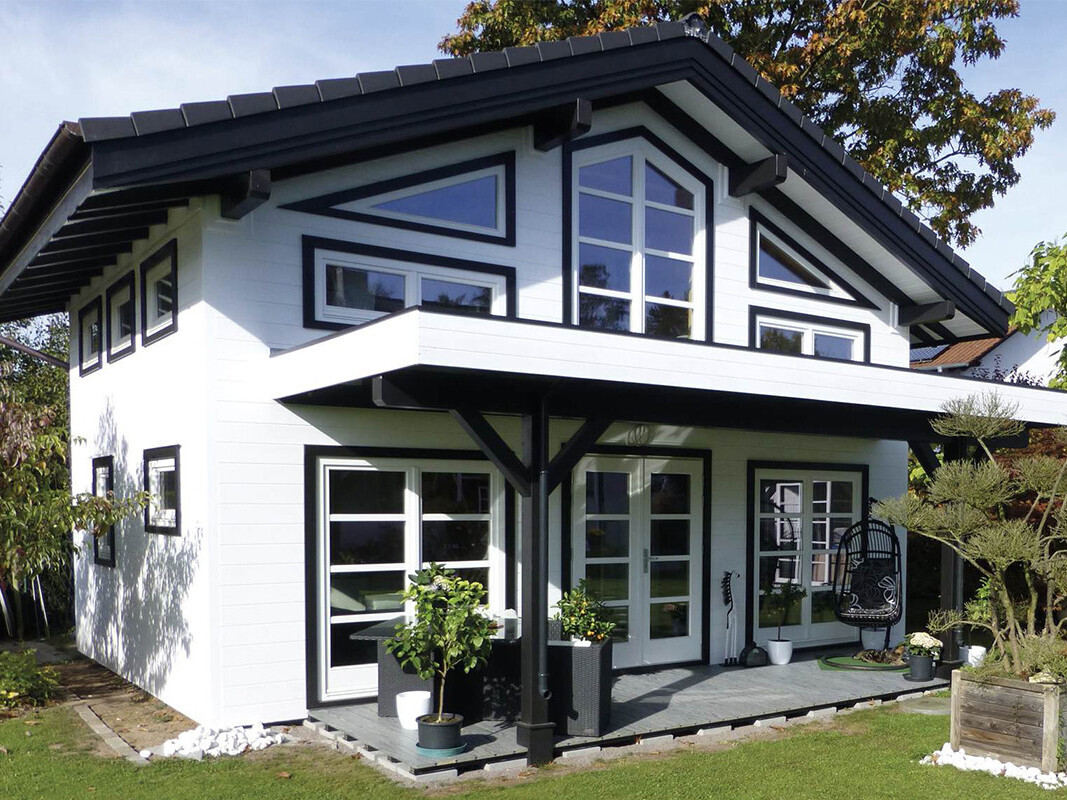 Elegant two-story timber house with a modern pitched roof and striking black and white design. The exterior features large triangular and rectangular windows framed in black, creating a contemporary and stylish look while maximizing natural light. A spacious covered veranda with black timber posts provides a comfortable outdoor seating area. The house is surrounded by a beautifully landscaped garden with lush greenery and decorative planters. Supplied by Lugarde and thoughtfully designed and installed by Forever Timber, this timber house seamlessly blends innovative design with exceptional craftsmanship for a luxurious and functional living space.