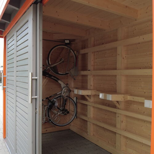 Interior view of a modern timber storage building showcasing neatly organized wooden shelving and wall-mounted bike racks, designed for efficient use of space. The natural wood interior contrasts with the sleek gray sliding doors, which provide secure and easy access. The bright orange frame adds a contemporary touch to the structure. Built on a sturdy paved surface, this versatile storage solution is perfect for organizing tools, bicycles, and other equipment. Designed and installed by Forever Timber and supplied by Bertsch HolzBau, it combines practicality, durability, and innovative design.