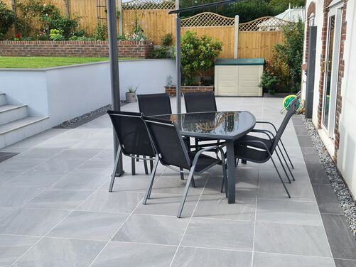 Stylish outdoor patio with large modern grey tiles, complemented by sleek black garden furniture. The multi-tiered design features white retaining walls and neatly landscaped greenery, creating a functional yet elegant space for relaxation and entertaining