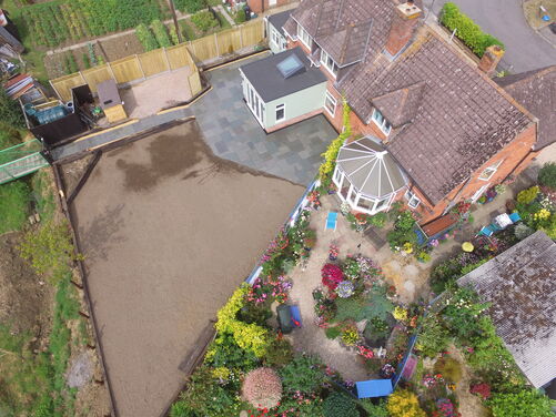 This aerial view showcases a property with a mix of established and developing outdoor spaces. On the right, there is a beautifully landscaped garden filled with vibrant flowers, greenery, and a conservatory extending from the house. The left side of the image reveals a more functional area under construction, featuring fresh soil and structural timber edging, likely for a new garden, lawn, or patio project.

The contrast between the lush garden and the under-construction section creates a sense of potential and transformation.