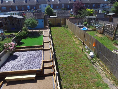 The image shows a garden that appears to be divided into two sections. On the left, there is a landscaped area featuring a small patch of artificial grass bordered by wooden sleepers, a section covered with decorative slate stones, and a pathway with a smooth gravel finish. A small tree and blooming rose bushes enhance the left-hand area. On the right, there is a larger patch of natural grass with some bare or uneven sections, bordered by wooden fencing. A clothesline and a small inflatable pool can also be seen. The garden backs onto a row of houses with visible sheds and additional greenery.