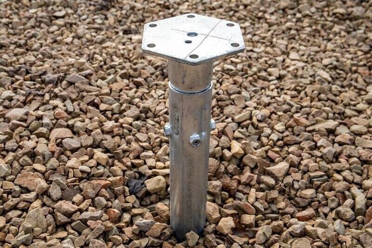 Close-up of a galvanized steel ground anchor embedded in a gravel base, designed for secure structural support. The anchor features a hexagonal mounting plate with pre-drilled holes for fastening and a sturdy cylindrical post with bolt fittings for stability. This durable component is ideal for foundations of outdoor structures, such as timber buildings, ensuring a reliable and long-lasting installation.