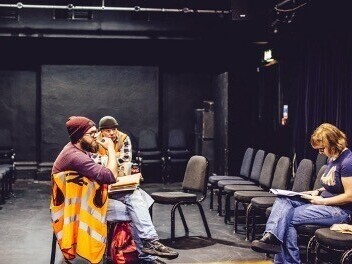 ​Robert ad Ted , wearing beanie hats and orange high vis jackets are sitting on either side of a table. Robert has a dark beard and glasses, he has his arm crooked, fingers resting on his lip. Ted is slightly bent towards the table with his arms crossed. They are both looking forward to the director who is sat on a chair facing them. She has a red bob, glasses and is wearing jeans and t-shirt. The director is focusing on her notes which she is adding to.