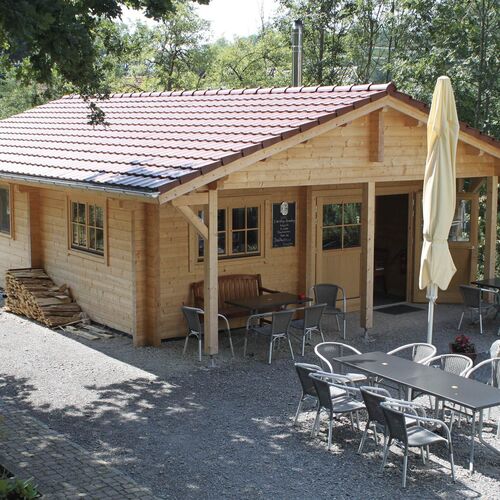 Rustic single-story timber house with a red-tiled pitched roof and a spacious covered porch. Constructed with natural wood, the structure includes charming wooden windows and a central entrance door. The outdoor seating area features multiple tables and chairs on a gravel surface, making it ideal for social gatherings or a cozy café setting. Stacks of neatly arranged firewood add a functional and decorative touch. Surrounded by greenery, this inviting space blends seamlessly with its natural surroundings. Supplied by Bertsch Holzbau and expertly designed and installed by Forever Timber, it combines practicality with a warm, welcoming ambiance.