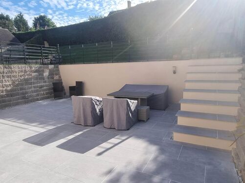 The image shows a well-designed, multi-tiered outdoor patio area. The lower level features a spacious patio with light gray tiles and several covered furniture pieces, including a table and chairs, providing a clean and low-maintenance look. A large potted plant adds greenery, softening the space.

To the right, a set of wide, modern steps leads to the upper level, bordered by retaining walls made of stone blocks. The upper tier is secured with sleek black railings and includes more greenery and functional outdoor space. Flower beds with vibrant plants are integrated into the retaining walls, adding color and natural beauty. The overall design combines functionality and aesthetics, making the space perfect for outdoor activities or relaxation.