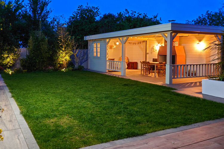 A well-lit backyard at dusk features a grass lawn and a bespoke wooden patio structure with a covered seating area. The patio, surrounded by lush greenery, has hanging lights and a dining set under the deep blue sky.