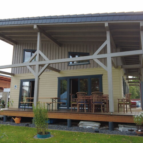 Contemporary two-story timber house featuring a large covered balcony and porch area, supported by decorative timber beams and braces. The exterior combines horizontal and vertical wood paneling in natural tones, complemented by dark-framed sliding glass doors and windows that allow ample natural light. The raised porch includes an inviting seating area with wooden tables and chairs, ideal for outdoor dining or relaxation. Set on a manicured lawn with landscaped elements, the house blends modern design with rustic charm. Designed and installed by Forever Timber, this home highlights exceptional craftsmanship and thoughtful architectural details.