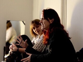 Lucy Hayward and director, Katnerine Wooton are sitting at a table. Lucy with shoulder length hair and a spotty shirt is focused on Katherine, who with long red hair snd a long sleeved t shirt has her arms raised as she excitedly describes the next steps.