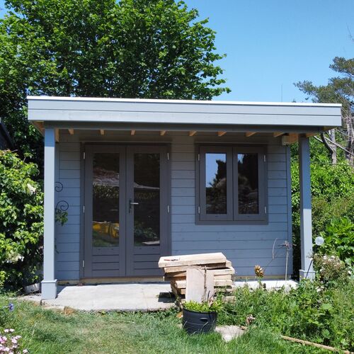 A stylish and practical wooden garden room designed and installed by Forever Timber, supplied by Lugarde. It features a soft grey exterior with a flat roof, a covered porch supported by two pillars, and a combination of double glass-panel doors and a side window for natural light. Surrounded by lush greenery, this versatile structure is perfect for a home office, garden studio, or peaceful retreat.
