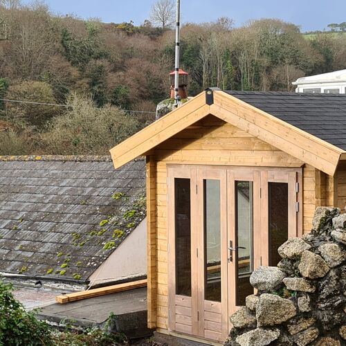 A cozy and rustic wooden garden building designed and installed by Forever Timber, supplied by Lugarde. It features a traditional pitched roof, natural wood finish, and double glass-panel doors for plenty of natural light. Nestled amidst a rural setting with stone walls and scenic greenery, this structure blends harmoniously with its surroundings, making it an ideal retreat or studio space.