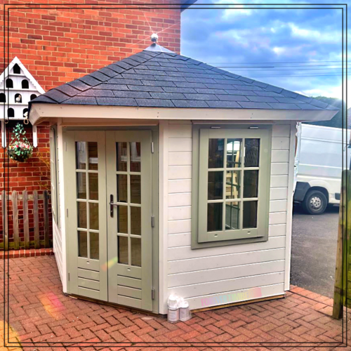 A bespoke timber garden building supplied by Lugarde and designed and installed by Forevertimber. This compact, hexagonal structure features a classic white-painted exterior with sage green-framed double glass doors and matching windows. The black shingle roof adds a traditional touch, while the design offers a balance of charm and functionality. Positioned on a brick-paved area next to a brick building, it serves as a stylish and practical addition, ideal for use as a garden retreat, hobby room, or cosy workspace. The design blends seamlessly with both modern and classic architectural styles.
