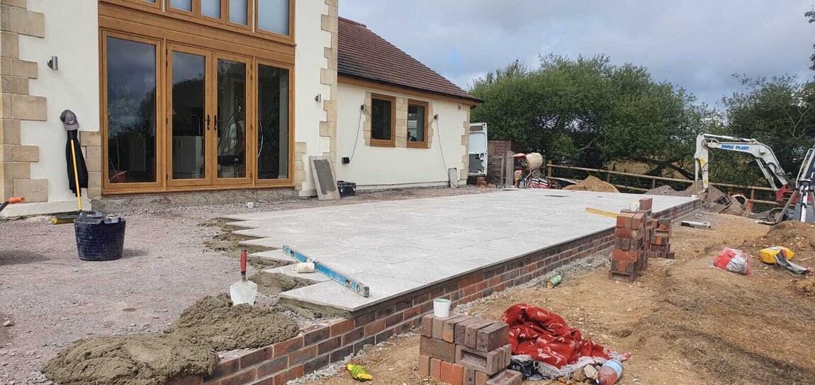 Construction progress of a stunning patio area with large stone tiles being laid on a raised brick base. The outdoor space complements the modern cream home with natural stone detailing and expansive wooden-framed glass doors, surrounded by lush greenery and scenic views.
