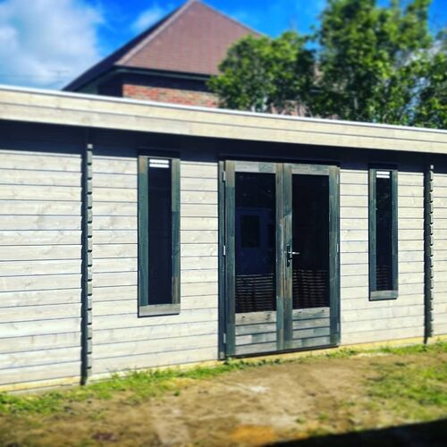 Modern timber garden building with a flat roof, featuring a sleek grey finish that adds a contemporary touch to its design. The structure includes double glass doors flanked by two tall vertical windows, providing ample natural light and a welcoming entryway. The clean lines and horizontal cladding give the building a minimalist aesthetic, making it a perfect addition to any modern garden. Ideal for use as a home office, studio, or recreational space, this building is expertly designed and installed by Forever Timber, ensuring both functionality and style.