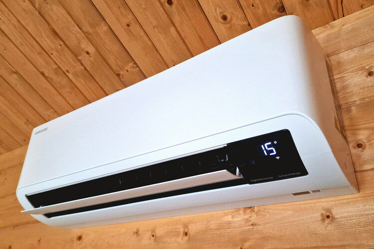 Close-up of a wall-mounted Samsung air conditioning unit installed against a timber-clad wall. The sleek, modern design features a digital display showing a temperature of 15°C, emphasizing precise climate control. The unit includes SmartThings integration for advanced functionality, offering both heating and cooling options. The natural wood paneling in the background highlights a warm, cozy setting, making this system ideal for maintaining comfort in residential or small commercial spaces