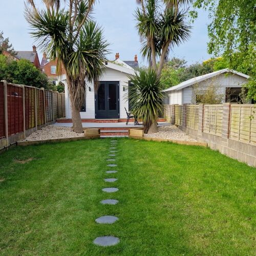 Charming timber garden building with a pitched roof, painted in a crisp white finish, and featuring dark-framed double doors and windows. The structure is situated at the end of a beautifully landscaped garden, with a lush green lawn bordered by decorative gravel beds and enclosed by wooden fencing for privacy. A stepping stone pathway leads to the building, flanked by tall palm trees, adding a tropical touch to the setting. This garden retreat, designed and installed by Forever Timber and supplied by Lugarde, serves as a stylish and functional addition, ideal for relaxation or as a versatile outdoor living space.