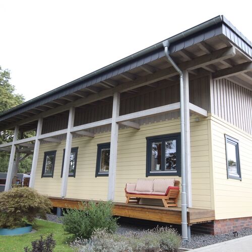 Contemporary single-story timber house with a flat-sloped roof, supported by exposed wooden beams that add structural elegance. The exterior features light beige siding with contrasting dark-framed windows, creating a modern yet inviting look. A raised wooden deck runs along the length of the house, complete with seating for outdoor relaxation. Set on a brick foundation and surrounded by neatly landscaped greenery, the house blends style with functionality. Supplied by Bertsch Holzbau and meticulously designed and installed by Forever Timber, this home offers a perfect balance of modern aesthetics and high-quality craftsmanship.
