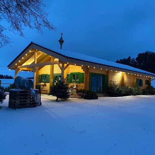 Cozy timber building illuminated beautifully at dusk, featuring a pitched roof with an overhang supported by exposed timber beams. The natural wood facade is complemented by green-framed windows with shutters, adding a charming rustic touch. String lights along the roofline enhance the inviting ambiance, while the surrounding snow-covered landscape creates a picturesque winter scene. A small outdoor area with festive evergreen trees adds to the seasonal charm. Designed and installed by Forever Timber, this structure combines warmth, craftsmanship, and functionality, making it perfect for residential or commercial purposes in all seasons.
