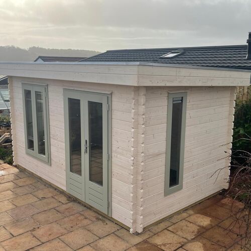 A bespoke timber garden building supplied by Lugarde and designed and installed by Forevertimber. This elegant structure features a flat roof and a light, natural wood finish painted in soft white tones. The building is accented with sage green-framed windows and double doors, which add a touch of charm and sophistication. The tall, narrow windows and glass-panelled doors provide plenty of natural light while maintaining a modern aesthetic. Set on a paved patio, this compact yet stylish building is ideal for use as a garden office, studio, or retreat space, blending functionality with contemporary design.