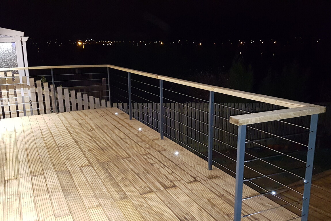 "Elegant timber deck illuminated by integrated LED lighting, featuring a sleek cable railing system with metal posts and wooden handrails. The elevated design offers stunning nighttime views of city lights in the distance, creating a cozy and modern outdoor space perfect for relaxation or entertaining.
