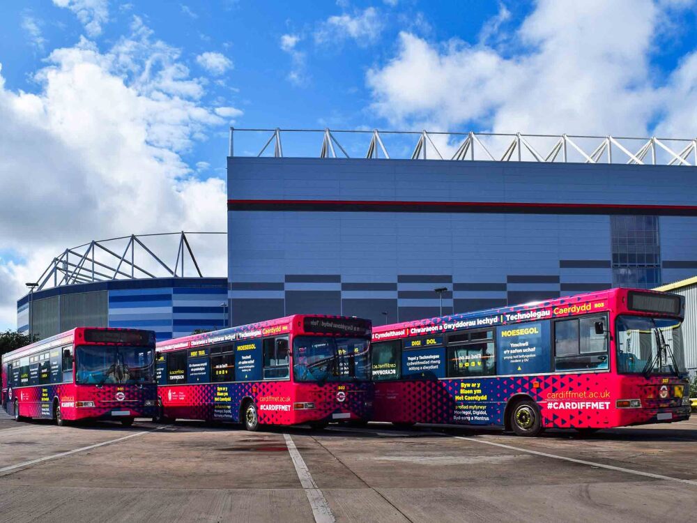 Fleet of Dennis Dart buses with matching Cardiff Metropolitan wraps and graphics parked outside football stadium.