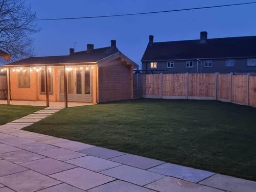 This nighttime image showcases a serene backyard setup with a wooden structure lit by warm string lights, creating a cozy ambiance. The pathway leading to the structure is neatly paved and divides a well-maintained lawn. The wooden fencing provides a sense of privacy and enclosure, while the distant glow from neighboring houses adds to the tranquil setting. The design is perfect for evening gatherings or quiet relaxation.