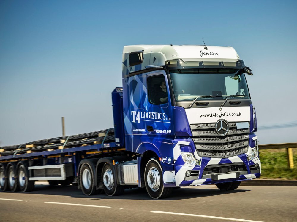Mercedes Actros driving along road with blue and white branded vehicle livery.