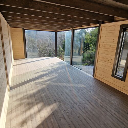 The interior of a bespoke timber garden building designed by Lugarde and crafted by Forevertimber. The space features a modern, open-plan layout with natural wooden paneling on the walls and ceiling, complemented by a matching wooden floor. Large sliding glass panels along one side flood the room with natural light, creating a bright and airy atmosphere. The exposed beams on the ceiling add a rustic yet contemporary charm, making it a versatile space suitable for a studio, office, or relaxation area.