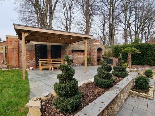 The image displays a beautifully landscaped outdoor entertainment area. A wooden pergola with string lights provides shade over a paved patio, creating a cozy seating space with a wooden bench. The patio is surrounded by carefully designed features, including a raised stone planter filled with neatly trimmed spiral-shaped shrubs, adding a formal and elegant touch to the space.

In the background, a red brick wall with decorative black panels and an arched brick structure houses an outdoor fireplace or barbecue area. The setting is framed by lush greenery, including hedges and a small palm tree, with tall, leafless trees towering in the background, creating a balance between formal landscaping and natural elements. This area is designed for both relaxation and entertaining.