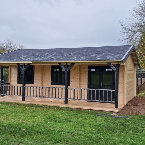 Charming single-story timber cabin with a pitched roof covered in durable shingles. The exterior showcases a natural wood finish, complemented by black-framed windows and doors for a striking contrast. A spacious covered porch runs along the front of the cabin, supported by decorative black timber posts and railings, offering a cozy outdoor seating area. Surrounded by a green lawn, this cabin is ideal for residential or recreational use. Designed and installed by Forever Timber, it blends rustic charm with modern craftsmanship to create a functional and visually appealing structure.
