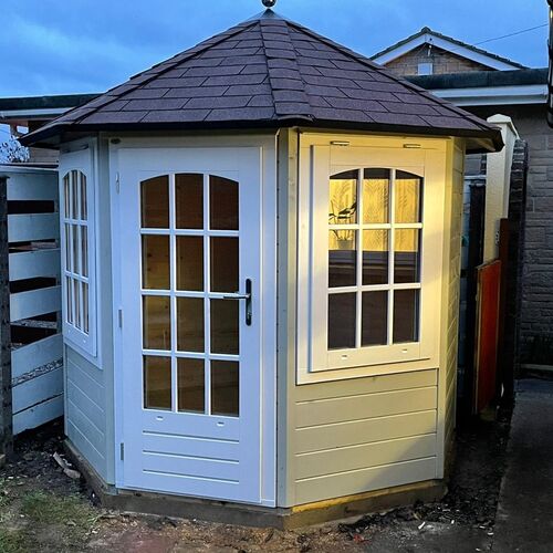 A charming hexagonal timber garden building supplied by Lugarde and designed and installed by Forevertimber. This compact structure features a light grey-painted exterior paired with white-framed glass-panelled double doors and windows, providing a timeless and elegant aesthetic. The arched window design adds character, while the dark brown shingle roof enhances its classic appeal. Warm lighting inside creates a cosy and inviting atmosphere, making it perfect as a garden retreat, reading nook, or decorative feature. Positioned in a compact space, this versatile building fits seamlessly into various garden settings.