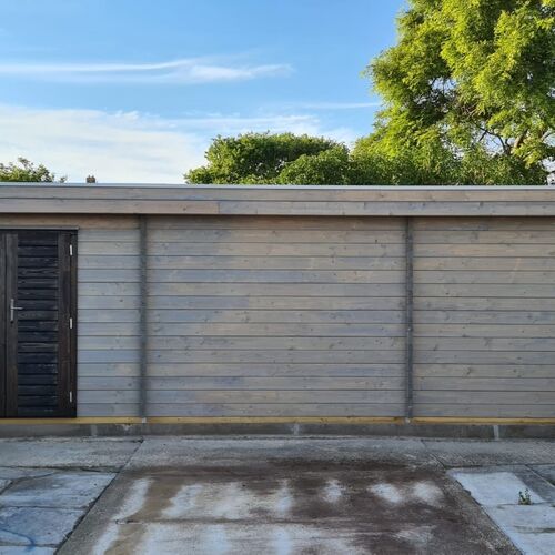 Spacious and modern timber garage with a flat roof, featuring a sleek horizontal plank design in a soft gray finish. Includes a contrasting dark-stained door with horizontal paneling and a steel handle, adding a bold and stylish touch. The garage is set on a robust concrete foundation with an extended paved driveway, ensuring durability and practicality. This custom garage, supplied by Lugarde and designed and installed by Forever Timber, combines contemporary aesthetics with exceptional craftsmanship for a functional and visually striking addition to any property.
