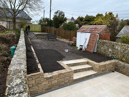 This image showcases a garden under development with freshly prepared soil and newly installed stonework. The steps and retaining walls made from natural stone blend beautifully with the existing stone wall and surrounding environment. The patio area with clean, light-colored tiles creates a seamless transition to the garden space. 

The small shed in the corner and other rustic elements add charm and character to the area, making it perfect for further landscaping or planting. If you're looking for ideas on how to enhance this space, such as adding seating, greenery, or functional features, feel free to ask!