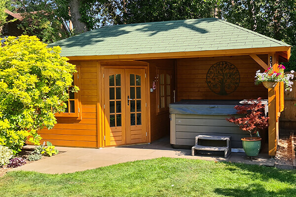 Charming timber garden building with a pitched roof covered in green shingles, featuring natural wood tones and a warm, inviting design. The structure includes double French doors with glass panels, a side window for additional light, and a sheltered open area perfect for outdoor relaxation. The open section houses a hot tub, adorned with decorative elements such as hanging flower baskets and a carved tree motif on the wooden wall. Surrounded by a well-maintained lawn and vibrant landscaping, this garden retreat is ideal for leisure and comfort in a tranquil outdoor setting.