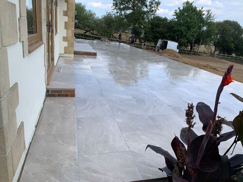 A modern house with a large, newly constructed patio featuring polished light grey tiles. The house has large glass windows and wooden-framed doors, with stone accents on the exterior. The surrounding area includes a landscaped garden with plants and an unfinished dirt section in the foreground