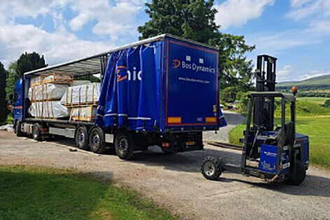 Delivery truck from Bos Dynamics parked in a rural setting, loaded with timber packages securely wrapped for transport. The truck features a blue curtain-sided trailer with company branding. A Moffett forklift is positioned next to the truck, ready for unloading materials. The scenic background includes greenery and rolling hills, indicating a countryside location. This image highlights the logistics and efficient delivery of construction materials for projects.