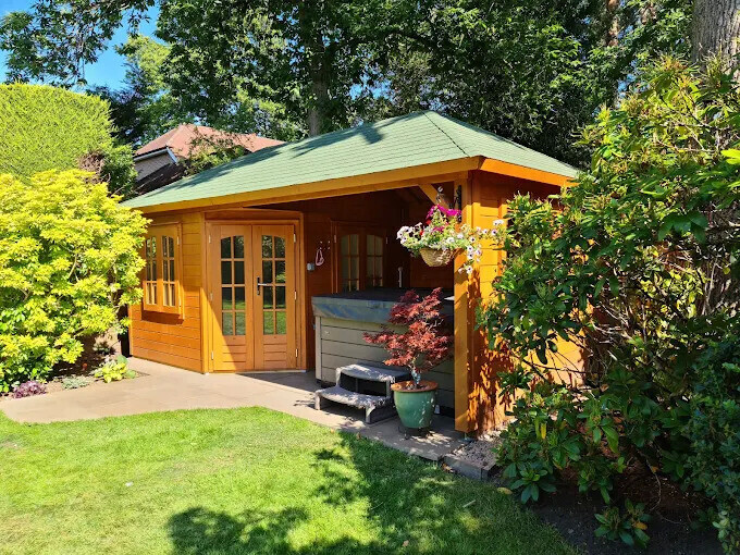 Charming timber garden building with a pitched roof covered in green shingles, featuring natural wood tones and a warm, inviting design. The structure includes double French doors with glass panels, a side window for additional light, and a sheltered open area perfect for outdoor relaxation. The open section houses a hot tub, adorned with decorative elements such as hanging flower baskets and a carved tree motif on the wooden wall. Surrounded by a well-maintained lawn and vibrant landscaping, this garden retreat is ideal for leisure and comfort in a tranquil outdoor setting.