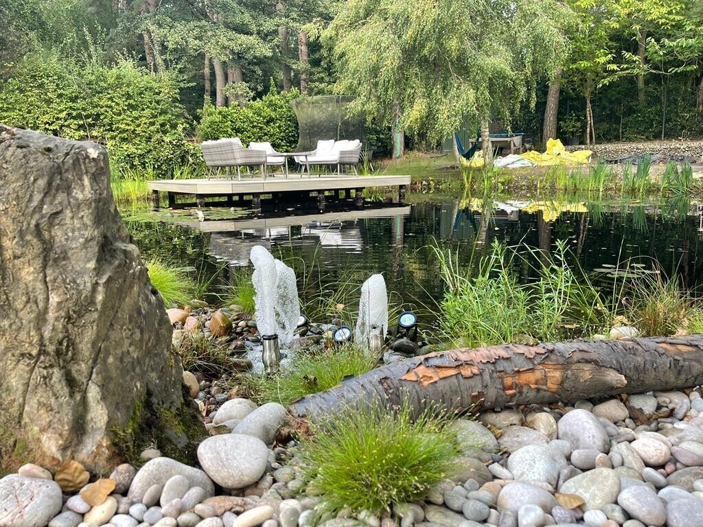 Tranquil outdoor seating area featuring a floating wooden deck over a serene pond adorned with lily pads. Surrounded by lush greenery and trees, this peaceful retreat offers a perfect setting for relaxation and enjoying nature.