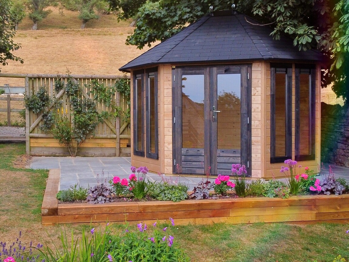 A charming timber garden building supplied by Lugarde and designed and installed by Forevertimber. This octagonal structure features natural wood paneling complemented by dark-stained frames on the full-length glass doors and windows, creating a striking contrast. The black shingle roof adds a classic touch, while the surrounding landscaped area includes vibrant flower beds bordered by wooden planters. This idyllic setup makes the building a perfect garden retreat or summerhouse, blending beautifully with the rural surroundings and offering a cozy, light-filled interior.